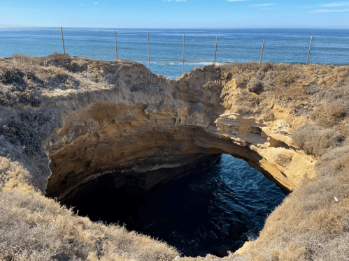 A deep, circular sinkhole near the ocean, surrounded by dry grass and a fence, with calm blue water below.