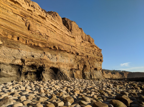Rocky coastline with layered cliffs and a beach covered in smooth, round stones under a clear blue sky.