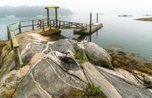 A foggy scene of a rocky shoreline with a small wooden dock and boats in the distance on calm water.