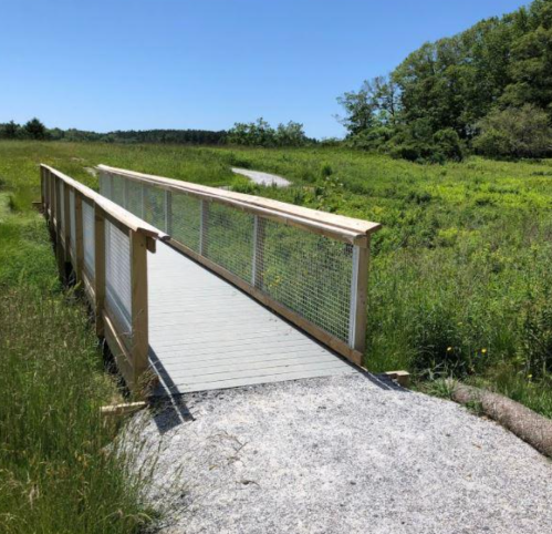 A wooden bridge spans a grassy area, leading into a lush green landscape under a clear blue sky.