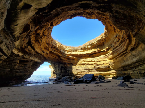 A sandy beach with a large rock arch, revealing a clear blue sky and calm ocean in the background.