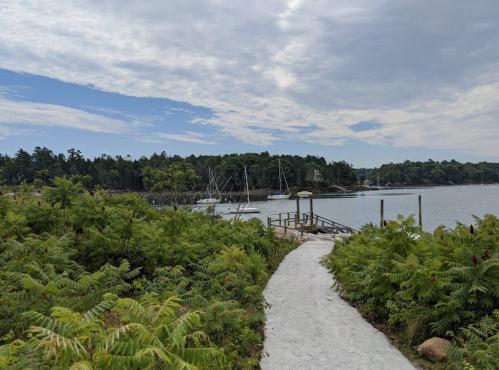 A sandy path leads through lush greenery to a calm bay with sailboats and a cloudy sky in the background.