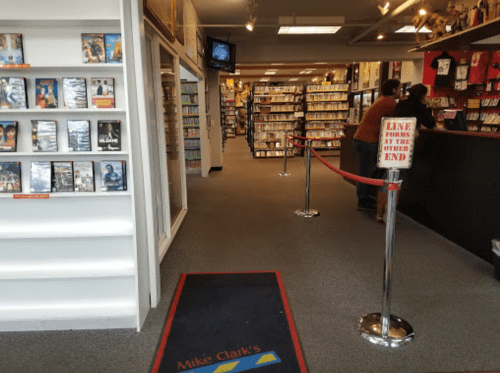 Interior of a video rental store with shelves of DVDs, a red rope barrier, and a sign directing customers to line up.