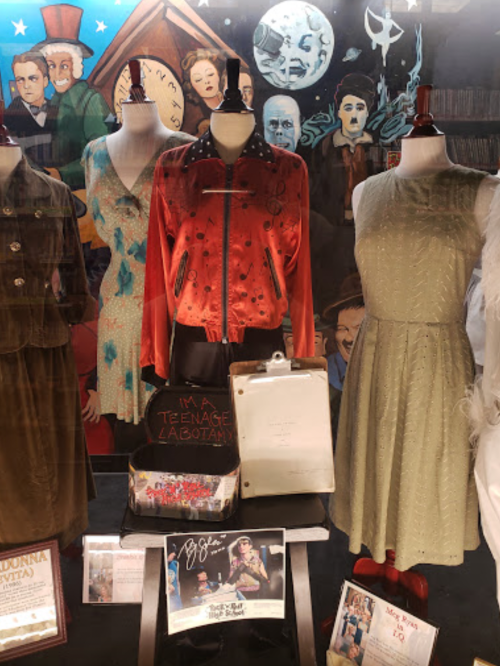 Display of costumes and memorabilia, including a red jacket and dresses, in a theatrical setting.