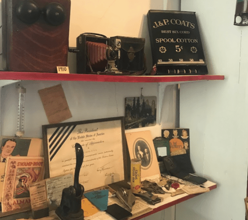 A display shelf featuring vintage items, including a camera, spool cotton sign, documents, and various antique tools.