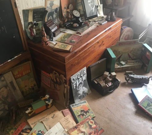 A cluttered room filled with vintage books, toys, and memorabilia scattered around a wooden chest and floor.