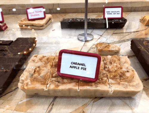 A display of fudge varieties, featuring a caramel apple pie fudge prominently labeled in front.