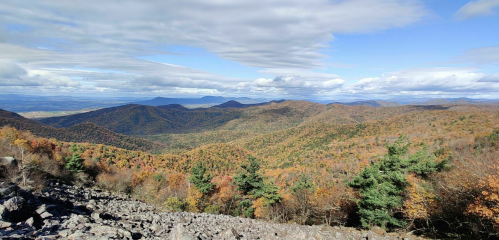 A panoramic view of rolling mountains covered in autumn foliage under a partly cloudy sky.