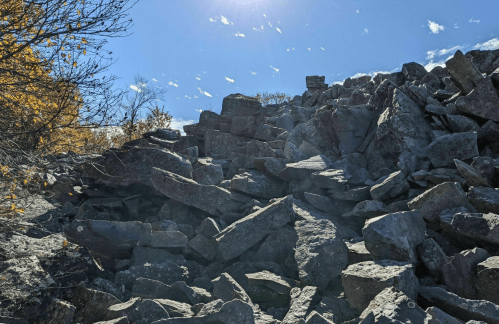 A rocky hillside under a clear blue sky, with scattered boulders and a few trees in the background.