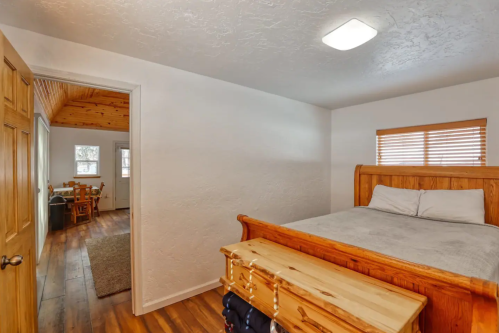 Cozy bedroom with a wooden bed and dresser, leading to a bright dining area through an open door.