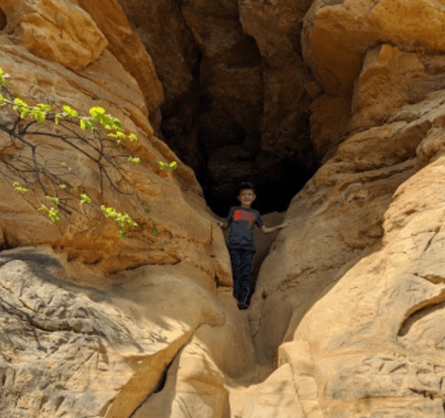 A child stands between two large rock formations, with greenery nearby and a cave-like opening in the background.