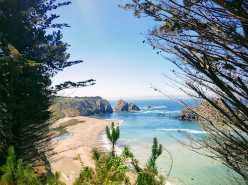 A scenic coastal view with rocky cliffs, sandy beach, and clear blue water framed by trees under a bright sky.
