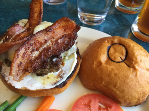 A close-up of a burger with bacon, cheese, and a fried egg on a bun, served with sliced tomatoes and carrots.