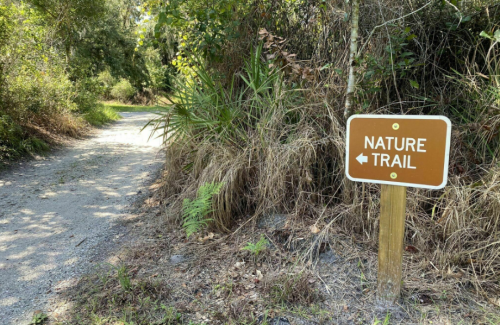 A brown sign points left to a nature trail, surrounded by greenery and a gravel path.