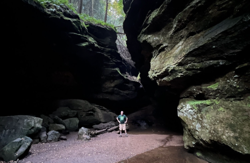 A person stands in a dark, rocky canyon surrounded by towering cliffs and lush greenery above.