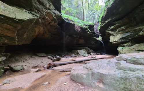 A serene cave with moss-covered rocks, a sandy floor, and small waterfalls surrounded by lush greenery.