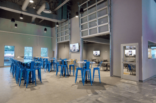 A spacious indoor area with blue chairs and tables, featuring large windows and screens on the walls.
