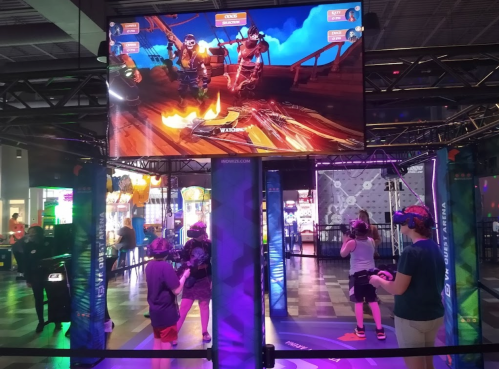 A group of people playing a virtual reality game in an arcade, with a large screen displaying the game action.
