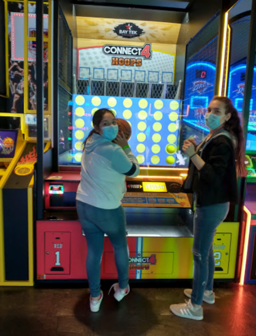 Two people wearing masks play a basketball arcade game called "Connect 4 Hoops" in a colorful gaming area.