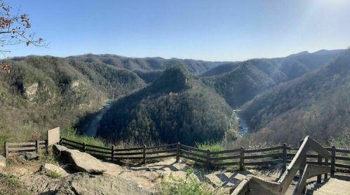 A panoramic view of rolling mountains and a winding river, with a wooden fence in the foreground under a clear blue sky.
