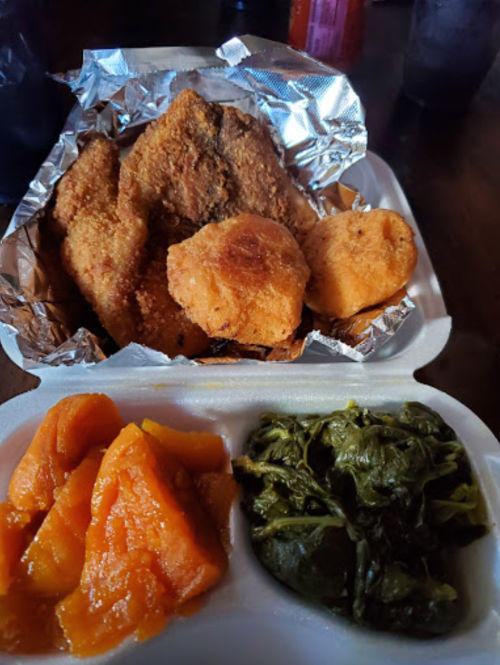 A takeout container with fried fish, cornbread, collard greens, and sweet potatoes.