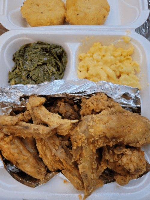 A takeout container with fried chicken wings, macaroni and cheese, collard greens, and two cornbread muffins.