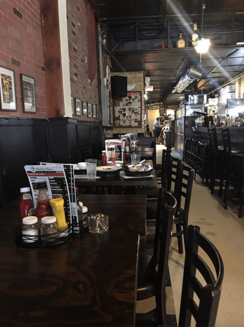 A cozy restaurant interior with wooden tables, black chairs, and condiment holders, featuring a brick wall and kitchen area.