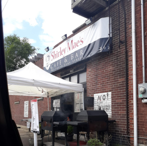 Sign for Shirley Mae's Cafe & Bar on a brick building, with a tent and outdoor grills visible.