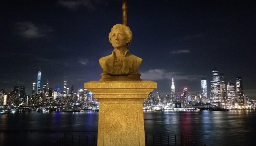 A bust of Alexander Hamilton overlooks a city skyline at night, reflecting lights on the water.