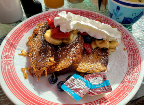A plate of French toast topped with bananas, strawberries, blueberries, and whipped cream, with butter packets on the side.