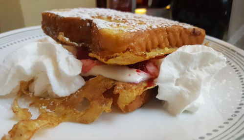 A plate with a stacked French toast sandwich filled with cream and strawberries, garnished with whipped cream and powdered sugar.