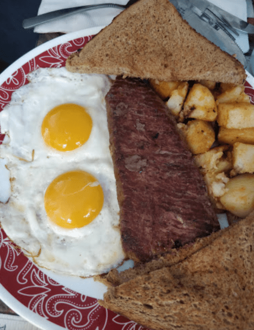 A plate with two sunny-side-up eggs, a slice of beef, roasted potatoes, and brown bread.
