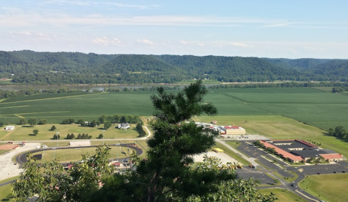 A scenic view from a hilltop overlooking green fields, a river, and a small town with buildings and roads.