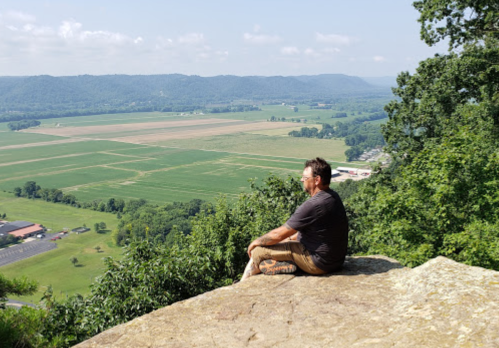 A person sits on a rock overlooking a lush green valley and distant hills on a sunny day.