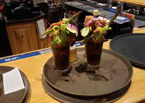 Two colorful cocktails garnished with vegetables, served on a tray at a bar.