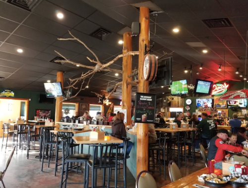 A lively restaurant interior with wooden decor, bar seating, and multiple TVs showing sports. Families and friends are dining.