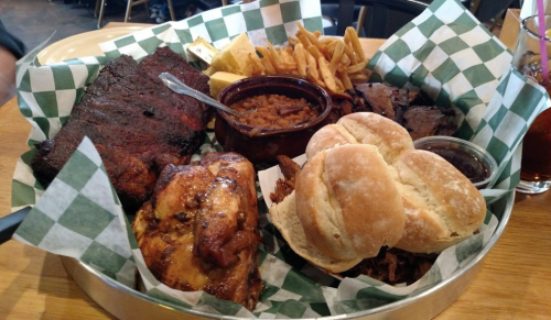 A platter of barbecue featuring ribs, chicken, pulled pork, baked beans, fries, and rolls, served in a basket.