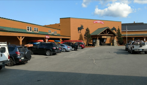 A rustic lodge with a wooden exterior, parked cars, and canoes outside under a clear blue sky.