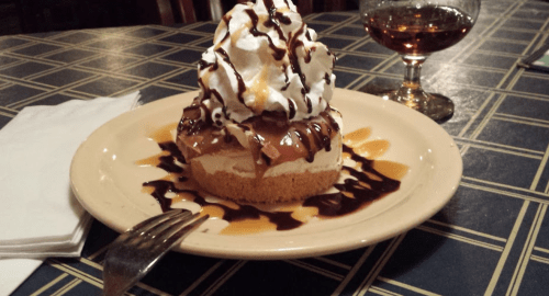 A slice of dessert topped with whipped cream and chocolate sauce, served on a plate with a glass of drink beside it.
