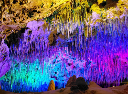 A colorful cave interior with vibrant stalactites illuminated in shades of purple, blue, and green.
