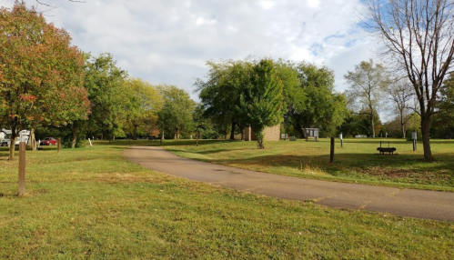 A winding path through a grassy park with trees and benches under a partly cloudy sky.