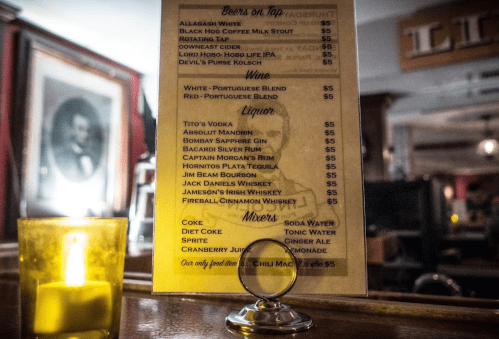 Menu board displaying drinks: beers, wine, liquor, and mixers, with prices listed. Candlelight in the foreground.