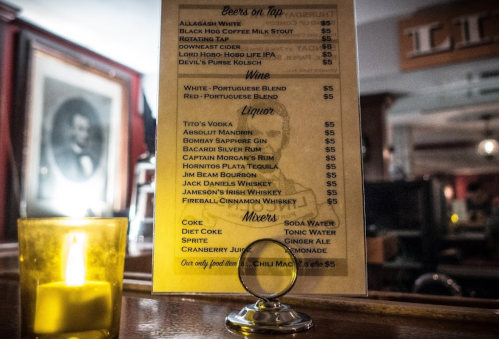 Menu board displaying drinks: beers, wine, liquor, and mixers, with prices listed. Candlelight in the foreground.