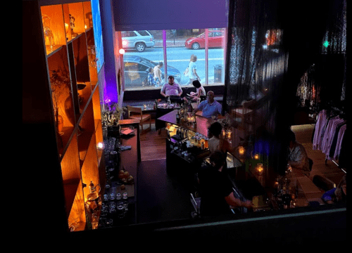 A cozy bar scene with dim lighting, shelves of bottles, and patrons enjoying drinks by a large window.