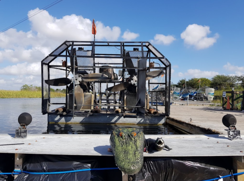 An airboat with large propellers is docked by a waterway, with a decorative alligator and raccoon figurine in the foreground.