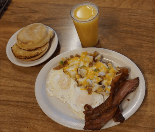 A plate of eggs, bacon, and cheesy hash browns, with a side of toasted biscuits and a glass of orange juice.