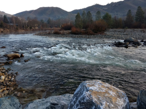 A serene river flows through a rocky landscape, surrounded by trees and mountains in the background.