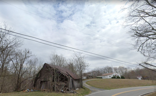 A dilapidated barn beside a winding road, with bare trees and a cloudy sky in the background.