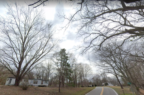 A rural road lined with bare trees, leading past an abandoned mobile home under a cloudy sky.