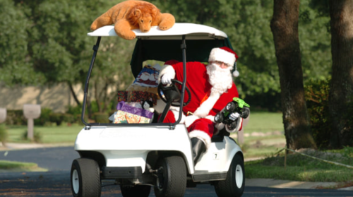 Santa Claus driving a golf cart, holding gifts, with a stuffed lion on top, in a festive outdoor setting.